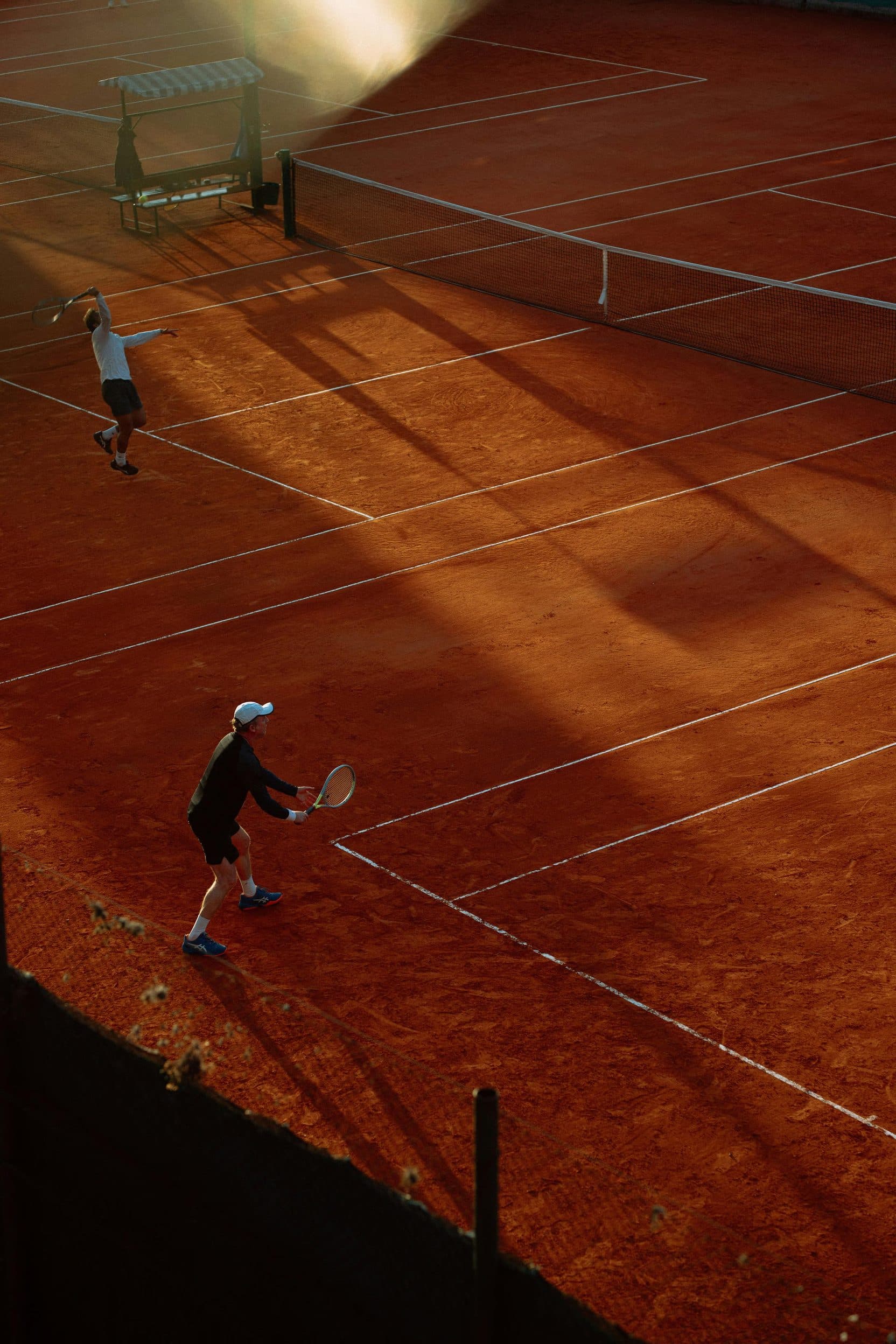 Two players on a terracotta clay court at golden hour, long shadows across the lines