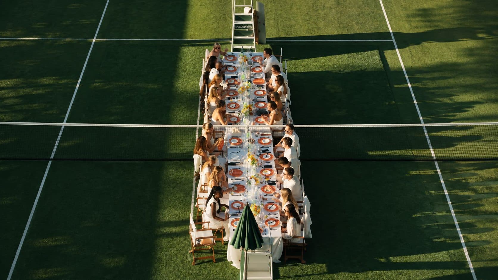 Guests in white at a long lunch table set along the centre line of a grass court